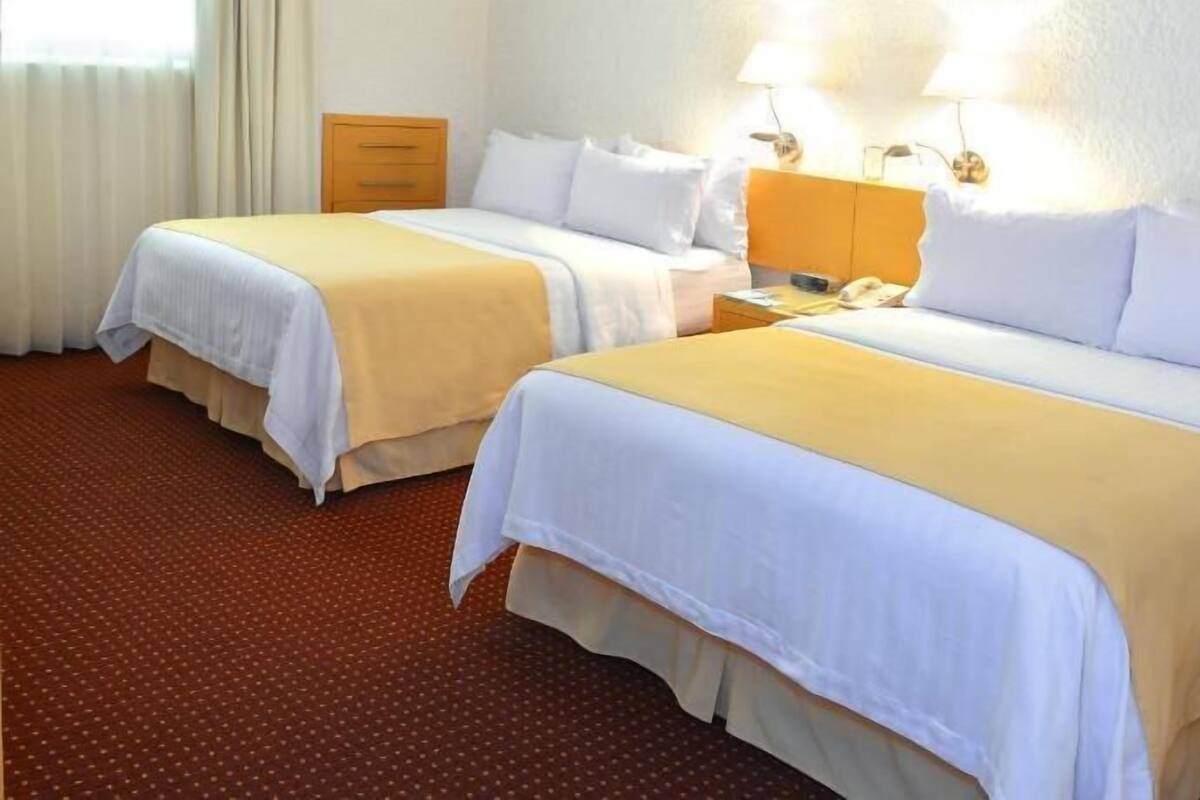 Une chambre d’hôtel avec deux lits, une table de chevet, une lampe et une fenêtre avec des rideaux.