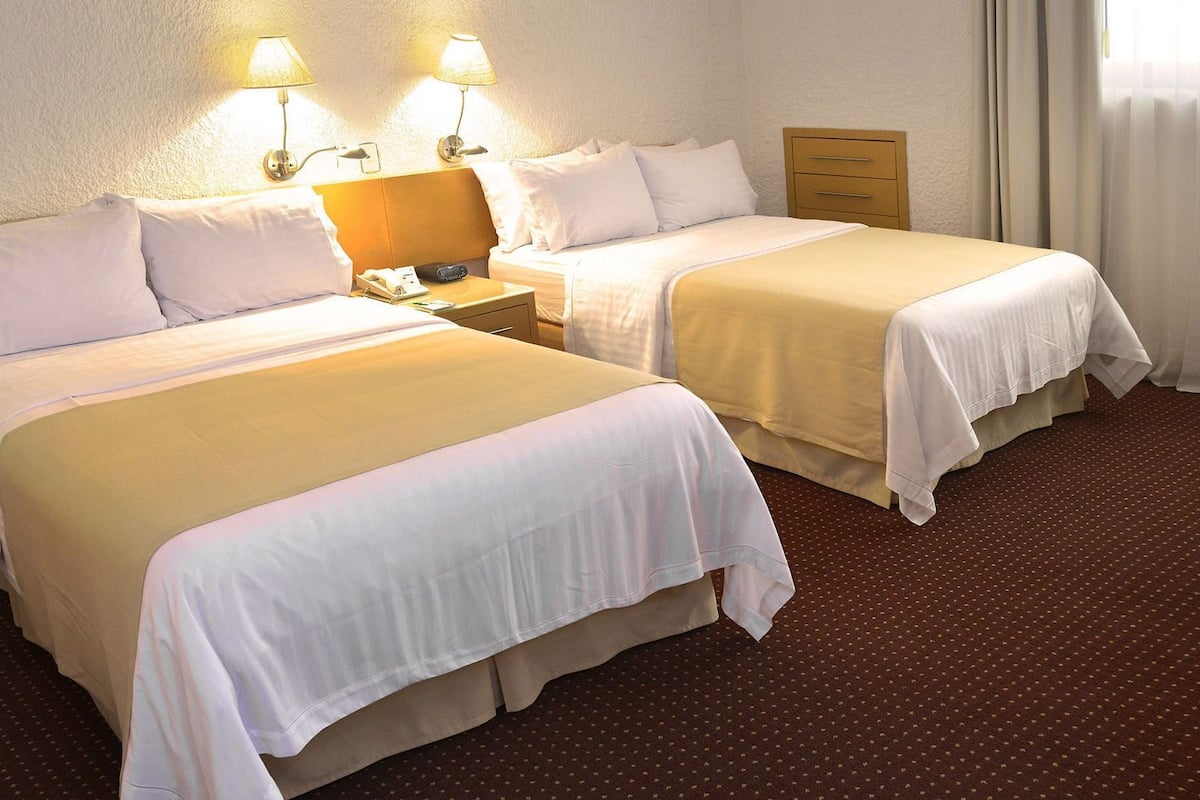 Une chambre d’hôtel avec deux lits, une table de chevet avec une lampe, une commode en bois et un sol recouvert de moquette.