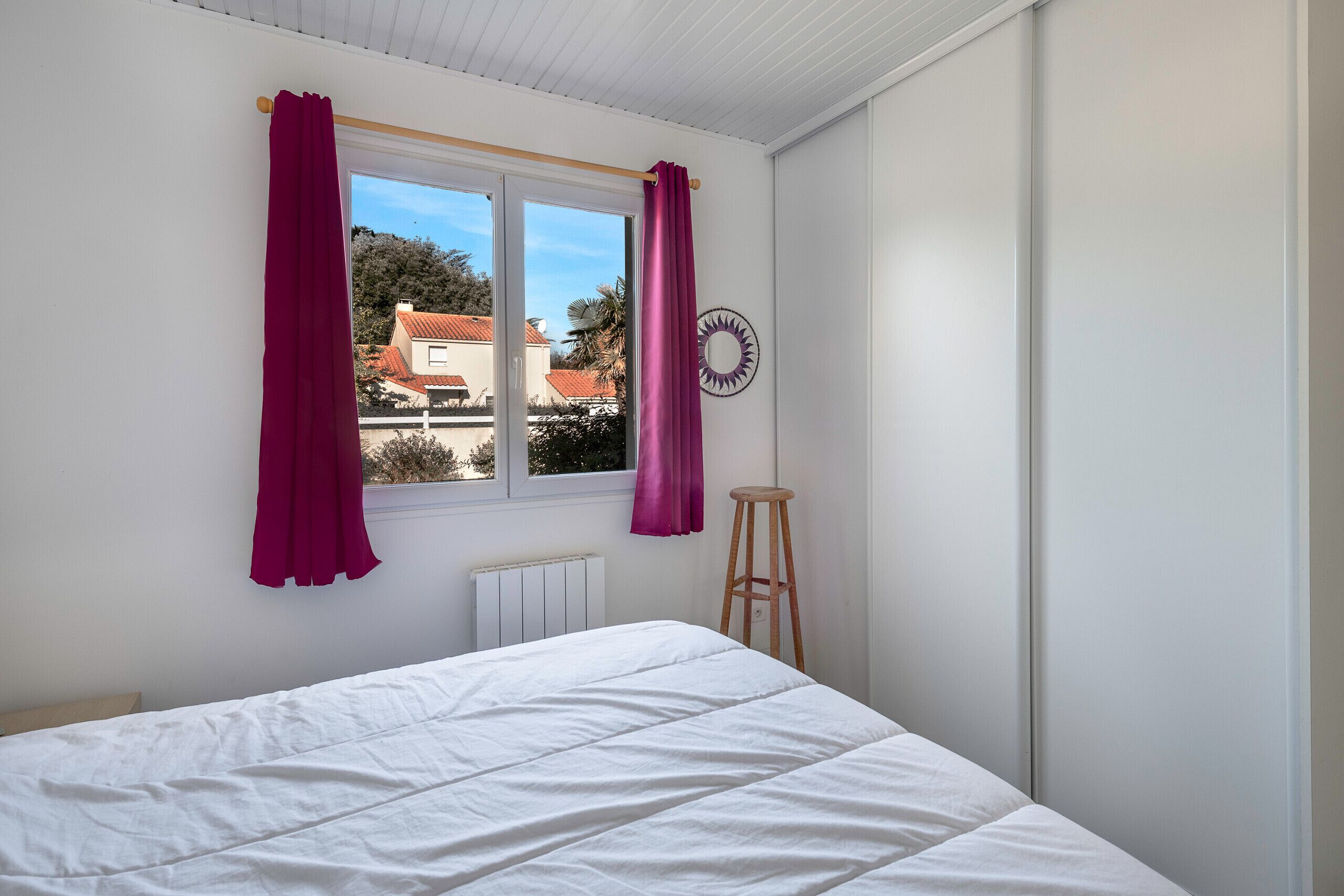 Une chambre à coucher avec un lit, une fenêtre avec des rideaux, un radiateur et un tabouret en bois.