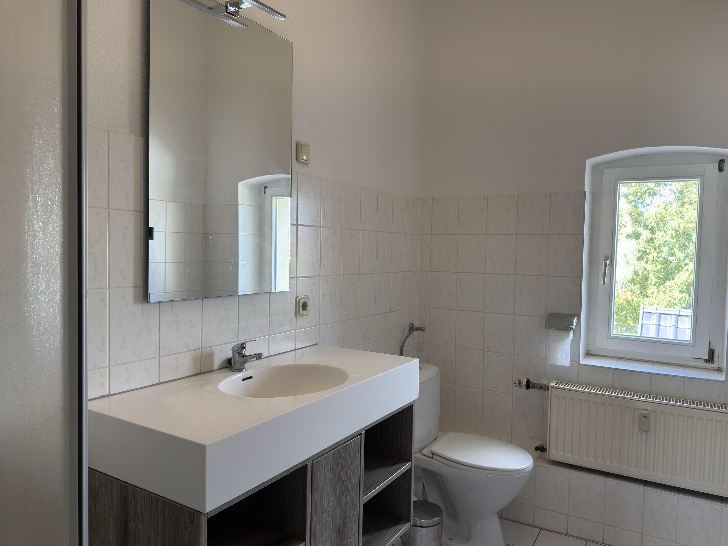 A bathroom with a white sink, mirror, and toilet. There is a window providing natural light.