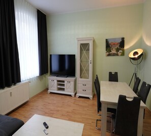 Un soggiorno con televisore, un mobile, un tavolo da pranzo con sedie, una lampada e un quadro appeso al muro.
