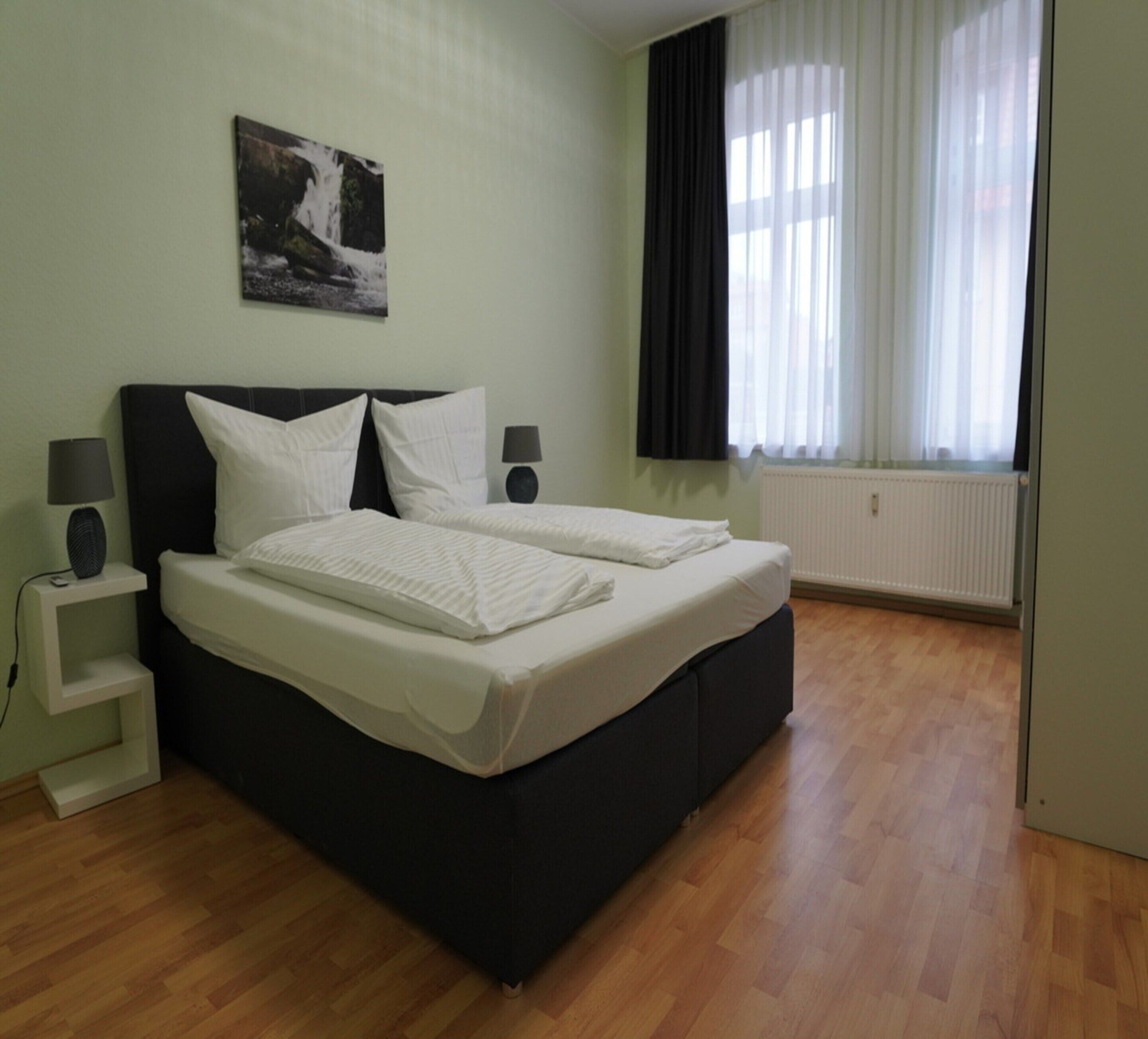 A bedroom with a bed, two bedside tables, a wall-mounted picture, a window with curtains, and a radiator.