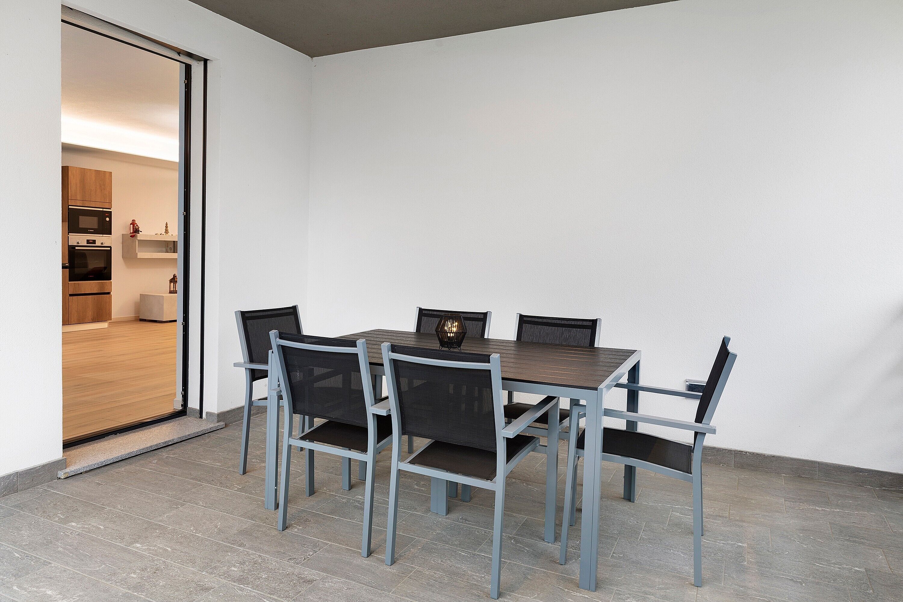A dining area with a table and chairs, a kitchen visible through an open door, and a tiled floor.
