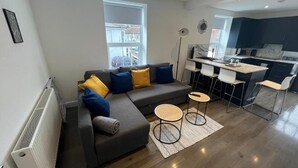 Una sala de estar moderna con un sofá seccional gris, dos mesitas redondas y vistas al área de la cocina.
