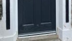 Una puerta delantera de color oscuro con un panel de vidrio y un tirador metálico, flanqueada por dos elementos decorativos negros.