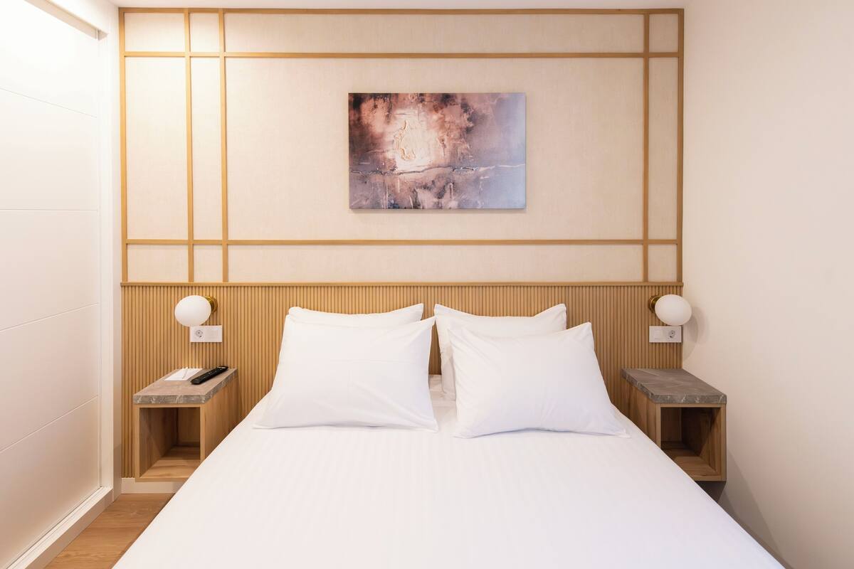 Une chambre d’hôtel comprenant un lit, deux tables de chevet, une petite table, une œuvre d’art accrochée au mur et une tête de lit en bois.