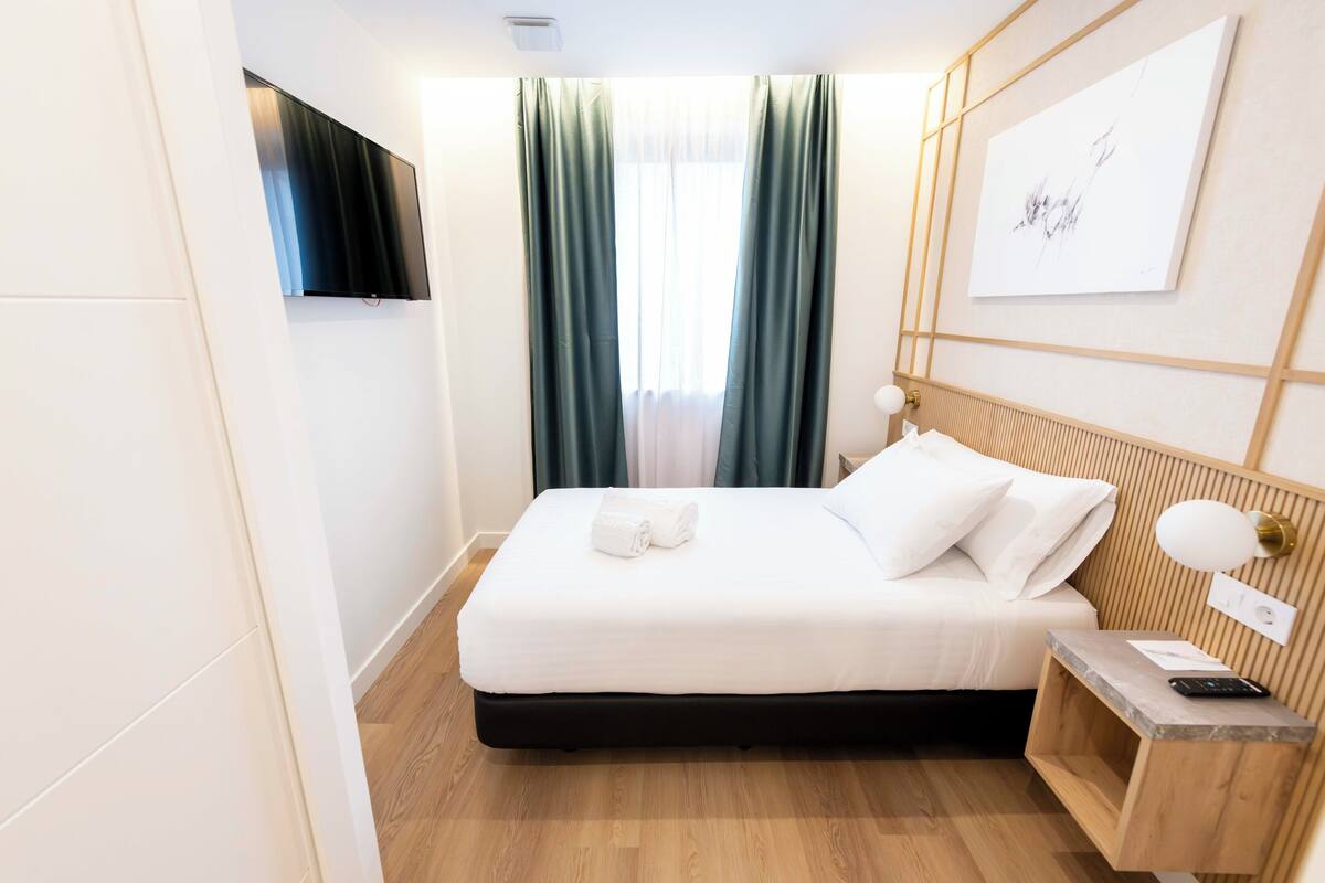 Un lit bien fait, avec du linge de lit blanc, une table de chevet avec une lampe et un téléviseur fixé au mur.