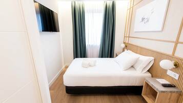 A neatly made bed with white linens, a bedside table with a lamp, and a wall-mounted TV.