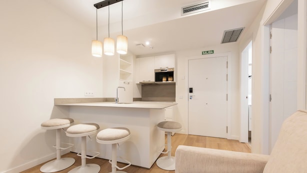 A modern kitchen with a bar area, white cabinetry, and a built-in oven. There are three stools at the bar and a sofa in the adjacent seating area. - Apartamentos Silken Goya (Oviedo)