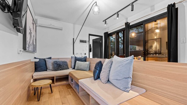 A modern living room with a wooden bench, a small round table, and a sofa with cushions. There is a large window with curtains, a ceiling fan, and a wall-mounted TV. - Stylish New Farm Apartment With Courtyard (Brisbane)