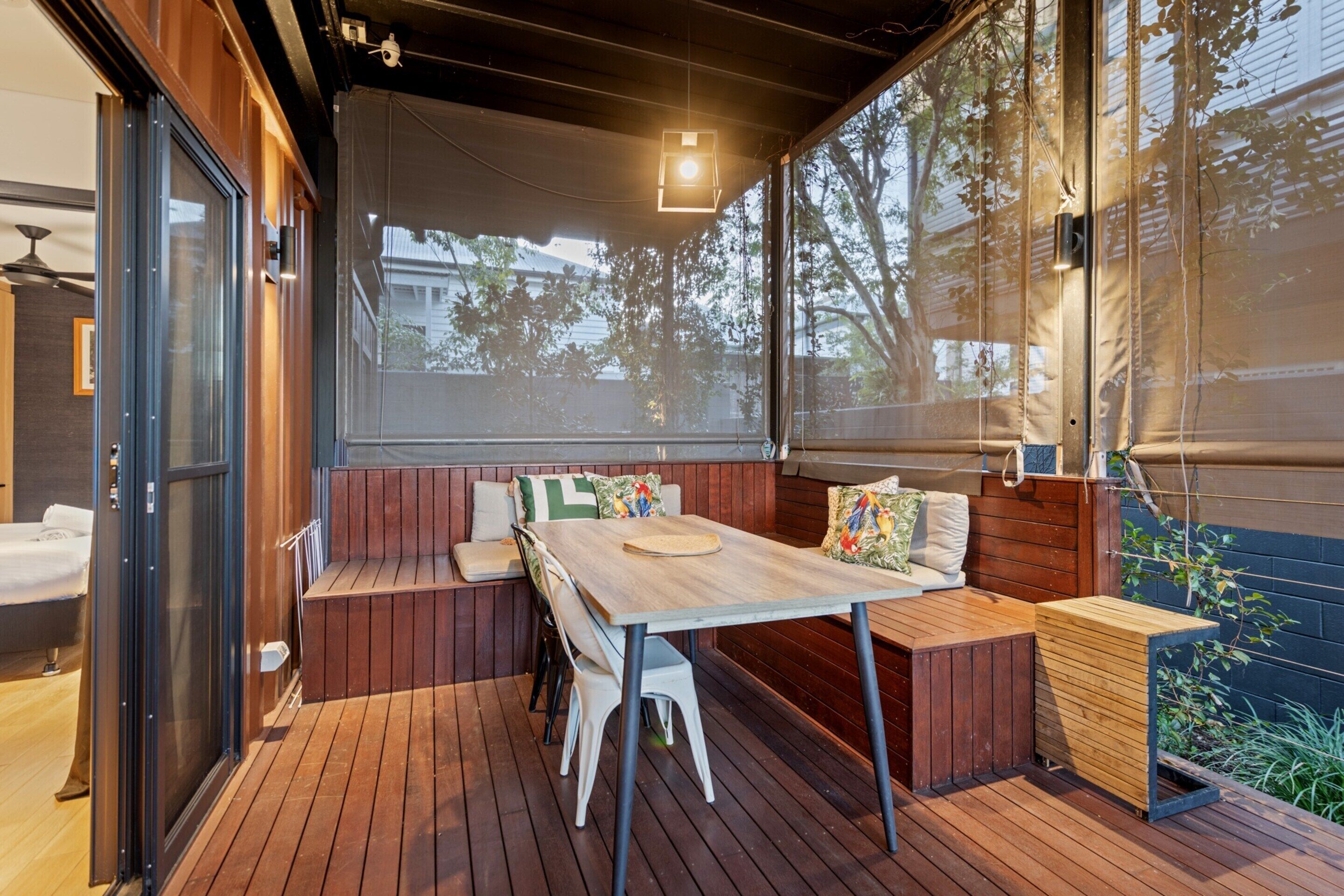 Un terrazzo in legno con tavolo e panche, una porta finestra e vista su alberi e palazzi.