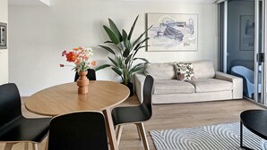 Un soggiorno moderno con un tavolo rotondo in legno, sedie nere, un divano beige con cuscini floreali, una grande pianta in vaso e un quadro astratto contemporaneo appeso alla parete.