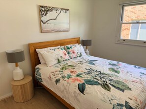 A bedroom with a wooden headboard, floral bedding, two bedside lamps, a round wooden nightstand, and a beach-themed wall art piece.