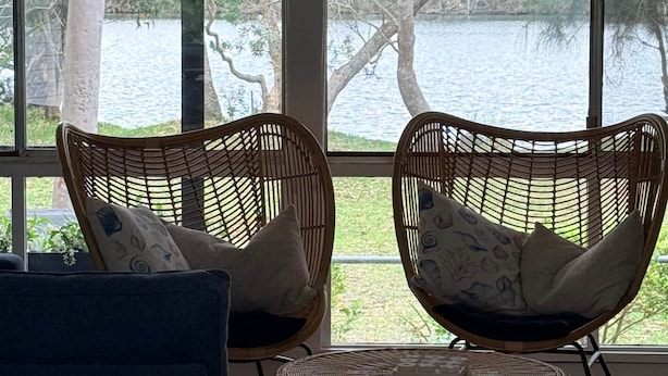 Une pièce avec deux fauteuils en osier placés face à une grande fenêtre donnant sur un plan d’eau et des arbres.