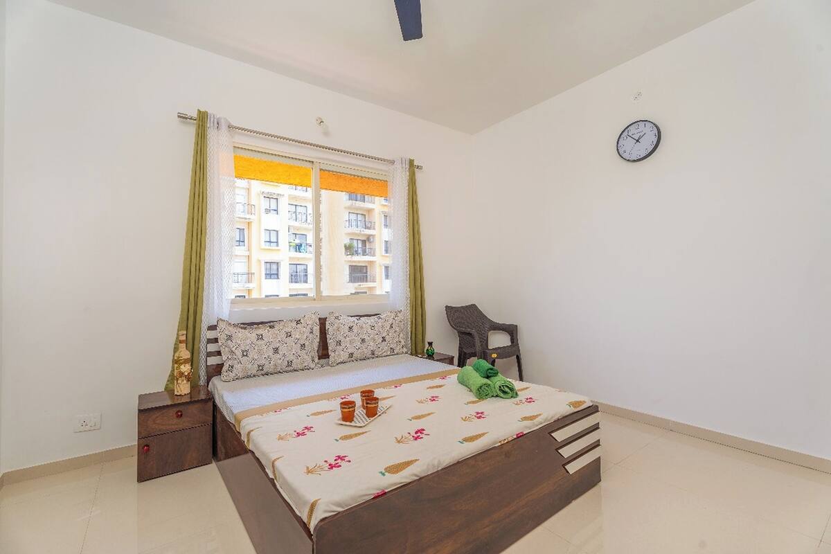 A bedroom with a bed, a nightstand, a chair, a window with curtains, and a wall clock.