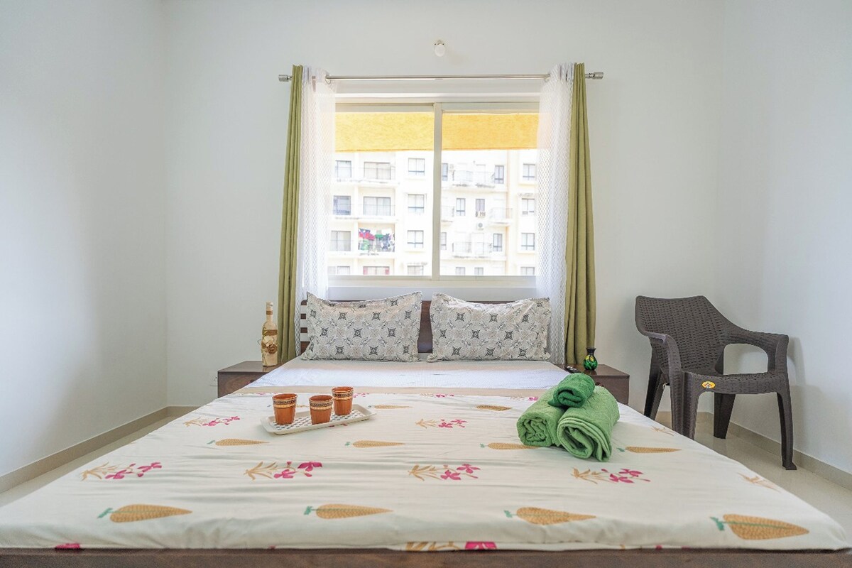 A neatly made bed with a floral bedspread, a small table with cups and a bottle, a green towel, and a chair.