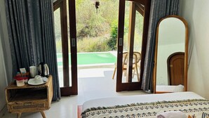 A bedroom with a bed, a wooden side table, a mirror, and a view of an outdoor area with a pool and greenery. - Felysia Villa III (Kintamani)