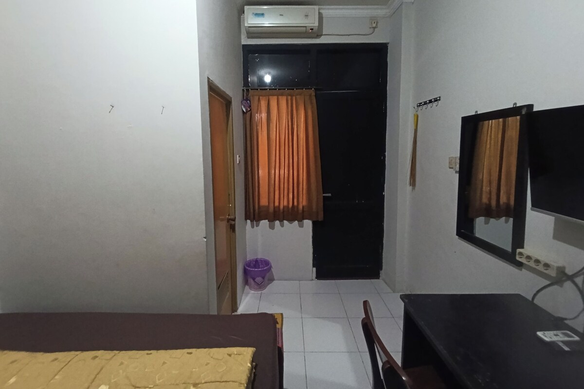 A small, dimly lit room with a bed, a desk, a chair, a window with curtains, and an air conditioning unit.