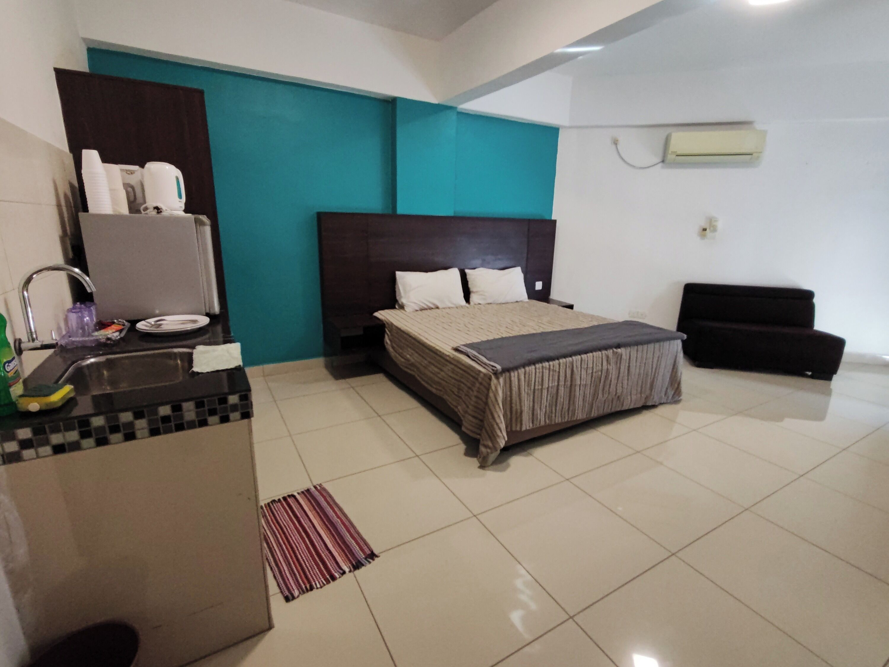 A hotel room with a bed, a small desk with a sink, a dark-colored sofa, and a teal accent wall.