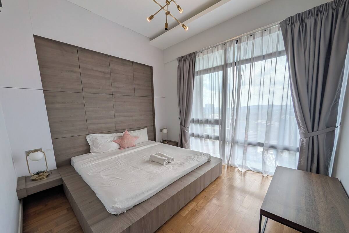 Ein modernes Schlafzimmer mit einem großen Bett, einem hölzernen Kopfteil, einem Nachttisch mit Lampe und Blick auf die Stadt durch leichte Vorhänge.