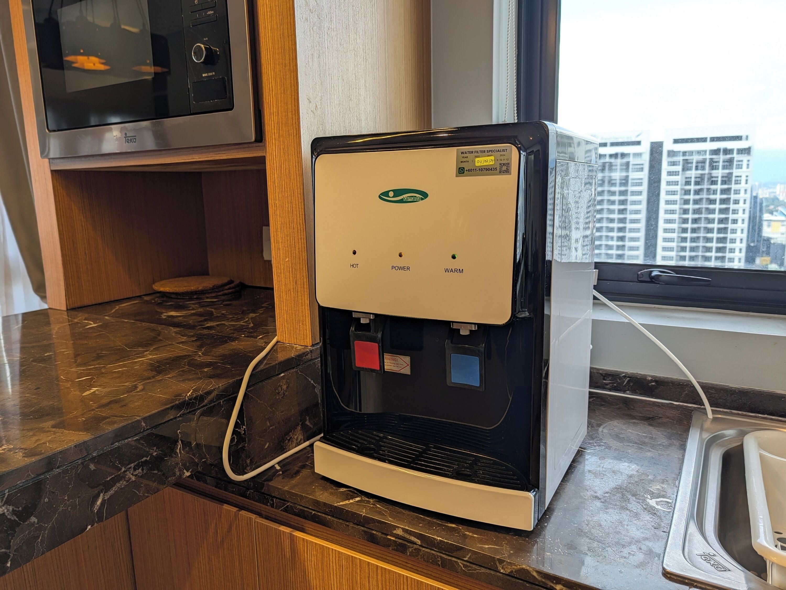 Een waterdispenser met warm, heet en lauw water, een magnetron en uitzicht op hoge gebouwen door een raam.