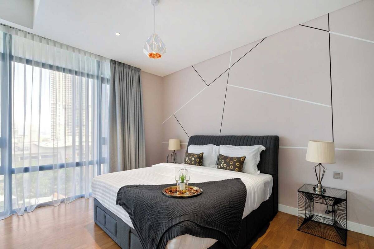 Ein modernes Schlafzimmer mit einem großen Bett, einem Nachttisch, einer Hängelampe und Blick auf die Stadt durch transparente Vorhänge.
