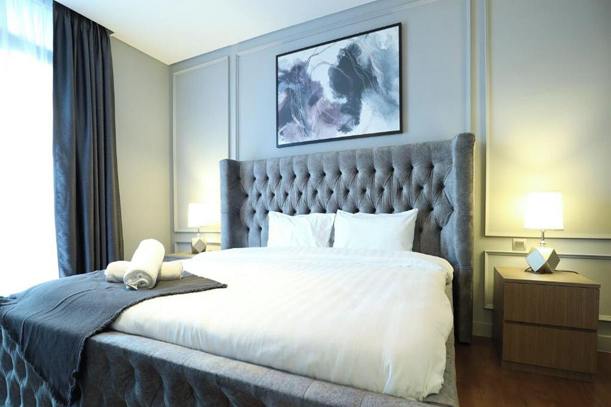Una cama grande con cabecera capitonada, dos mesitas de noche con lámparas, un cuadro abstracto enmarcado sobre la cama, y una ventana con cortinas.