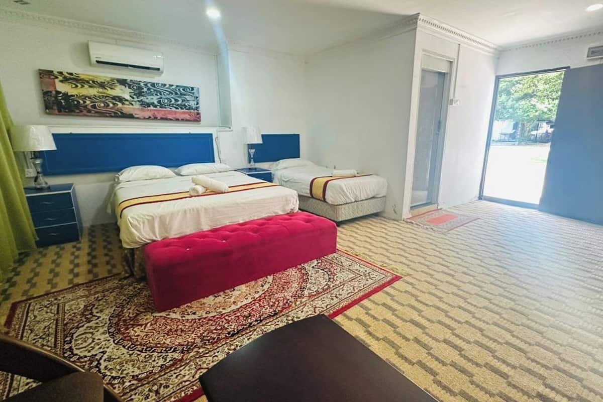 Une chambre d’hôtel avec deux lits, un pouf rouge, un tapis à motifs et une grande fenêtre.