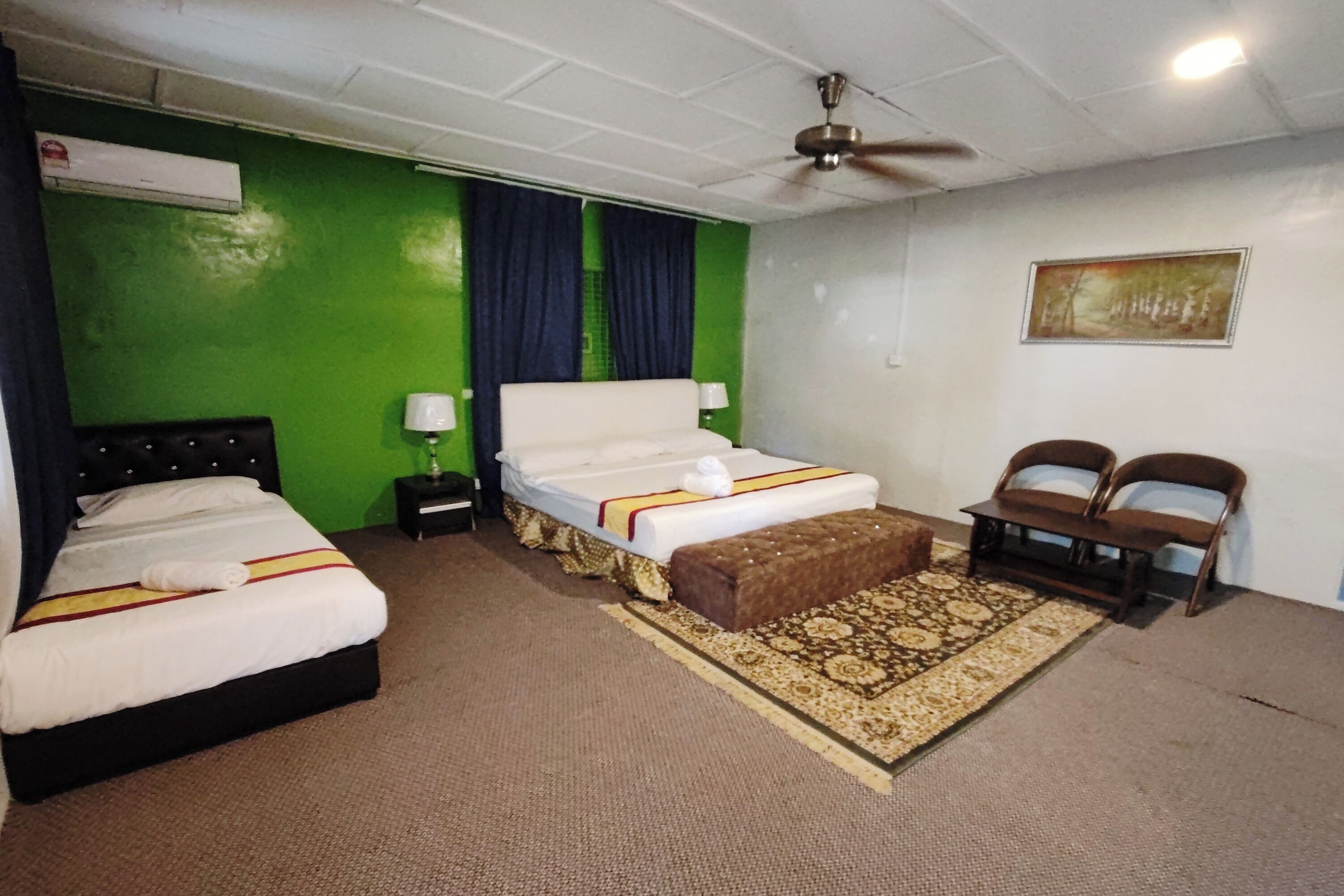 A hotel room with two beds, a ceiling fan, a green wall, a patterned rug, and a painting on the wall.