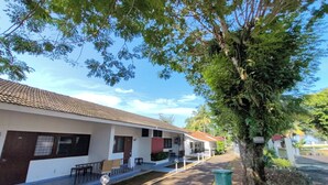 Un hilera de edificios de una planta con techo de tejas, mesas al aire libre y un árbol en primer plano.