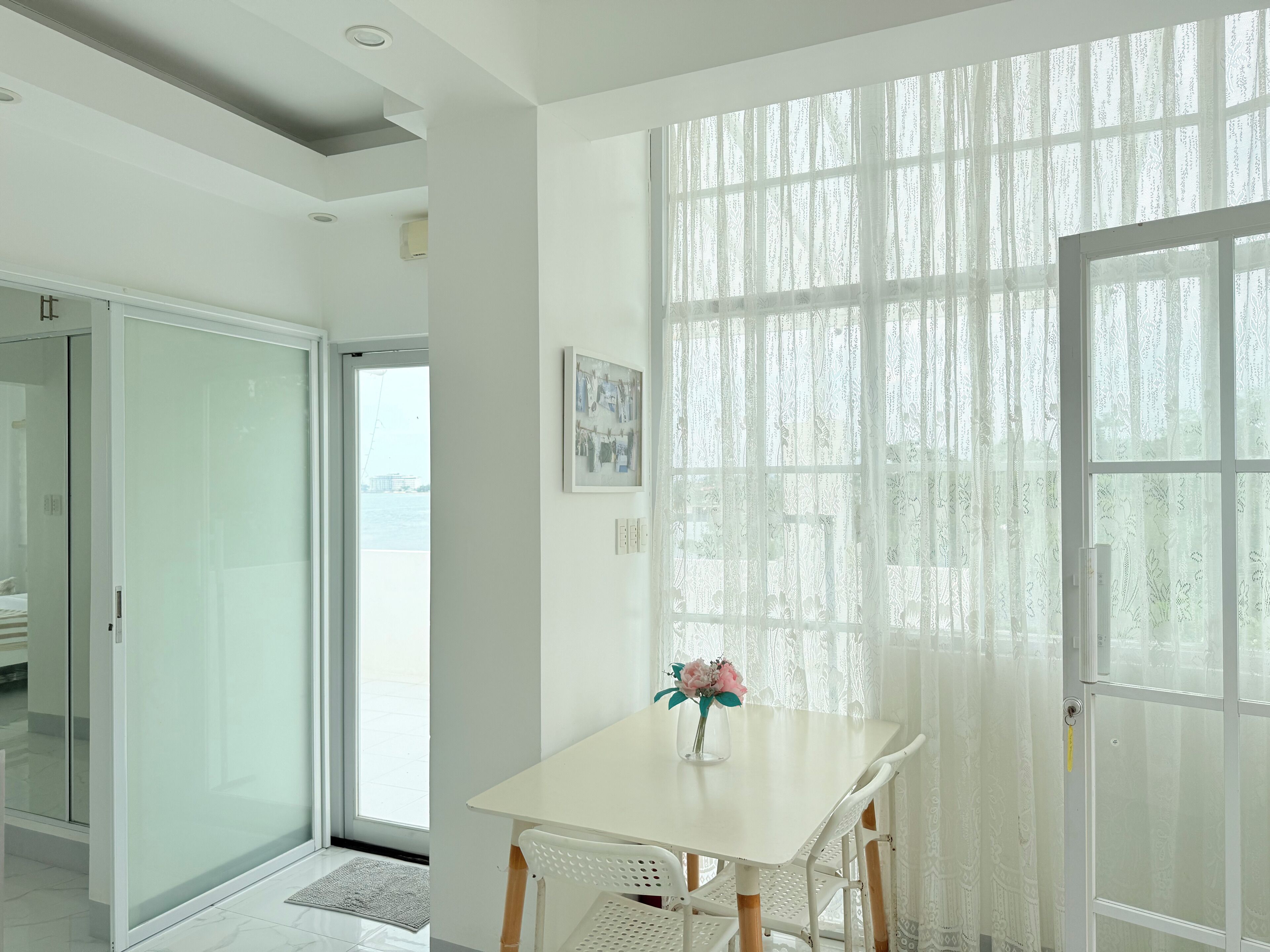 A modern room with a white table, a vase with flowers, and a view through sheer curtains.