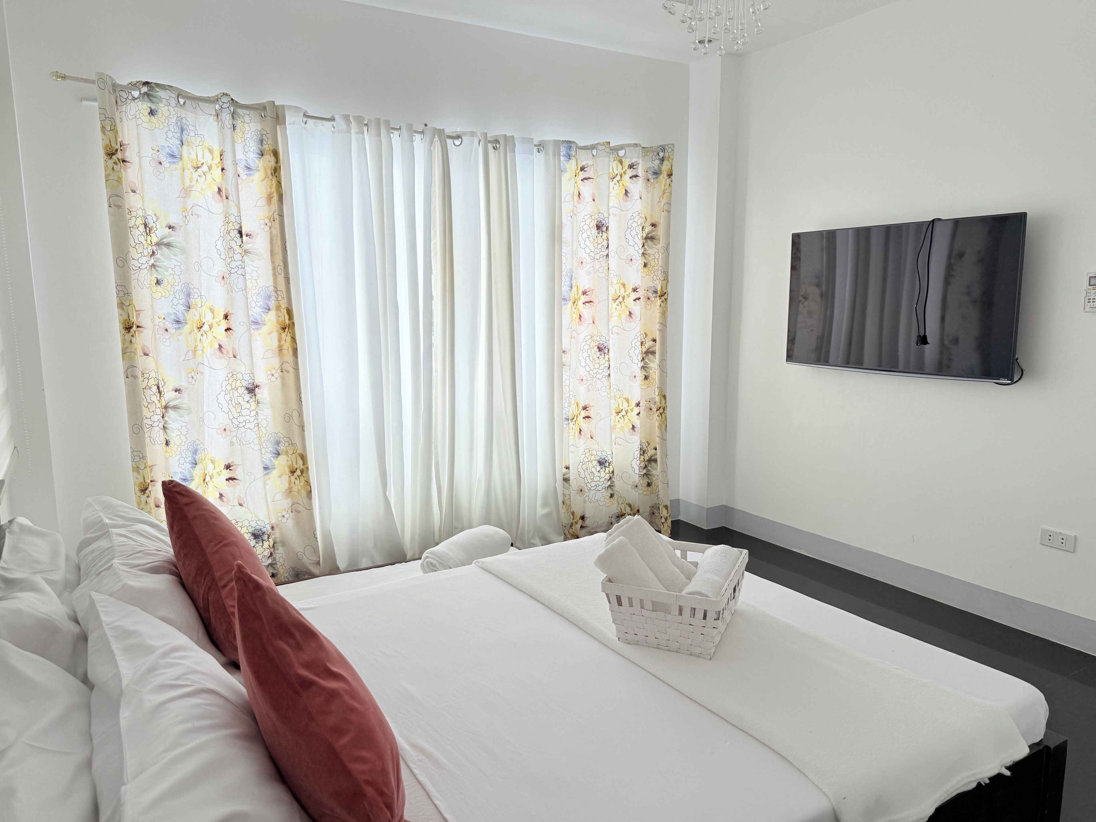 Habitación de hotel con cama, ropa de cama blanca, almohada roja, cesta blanca con toallas, cortinas con motivos florales y televisor de pantalla plana montado en la pared.