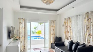 A modern living room with a black leather sofa, a white cabinet, and a large window with a view of a pool and greenery. - Chateau Du Soleil (San Fernando)