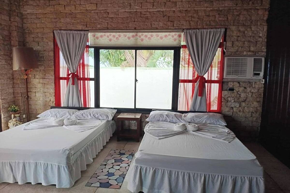 A room with two beds, a brick wall, a window with curtains, an air conditioning unit, and a small table with a lamp.