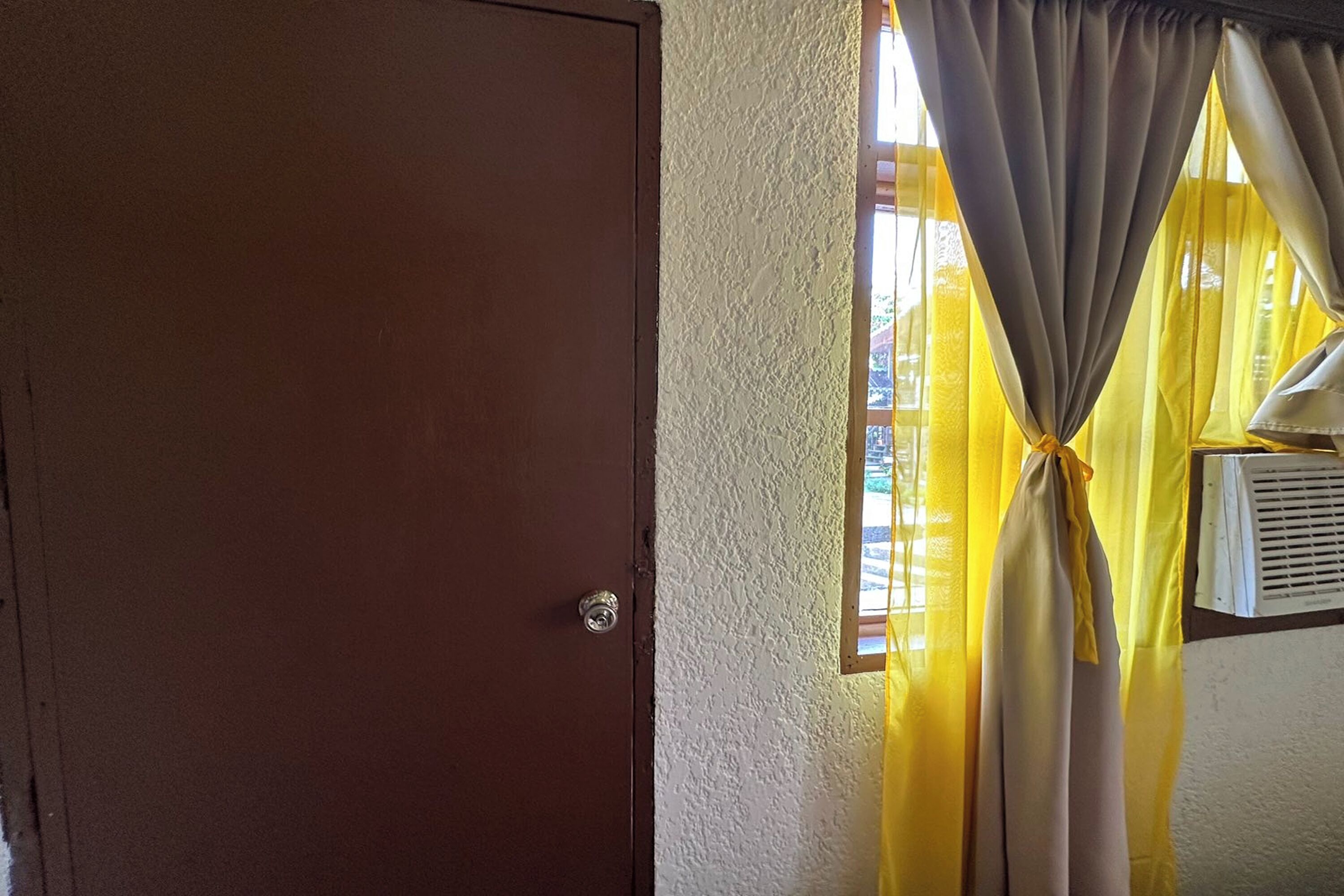 A room with a wooden door, a window with yellow curtains, and an air conditioning unit mounted on the wall.