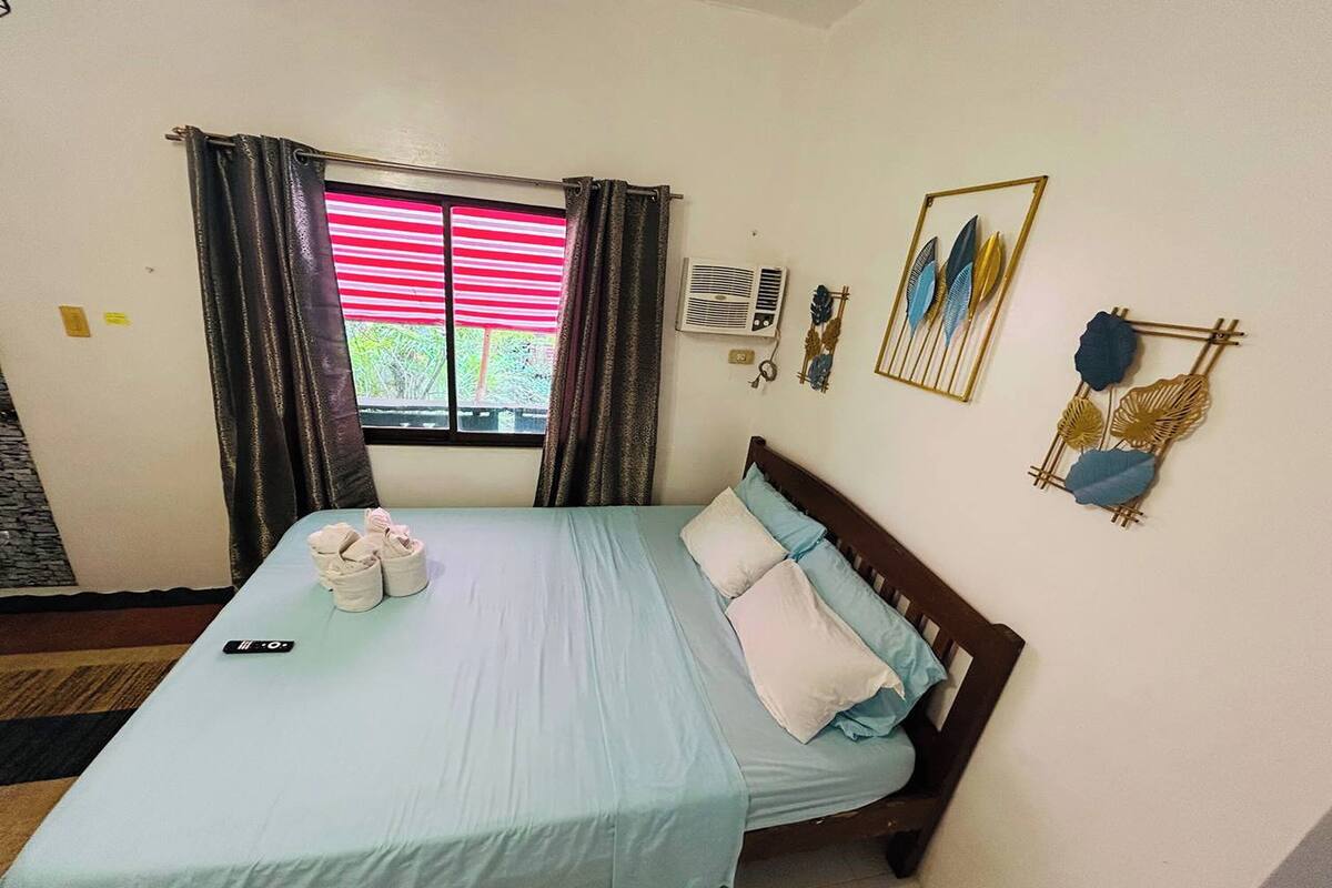 A hotel room with a bed, a window with curtains, a wall-mounted air conditioner, and decorative wall art.