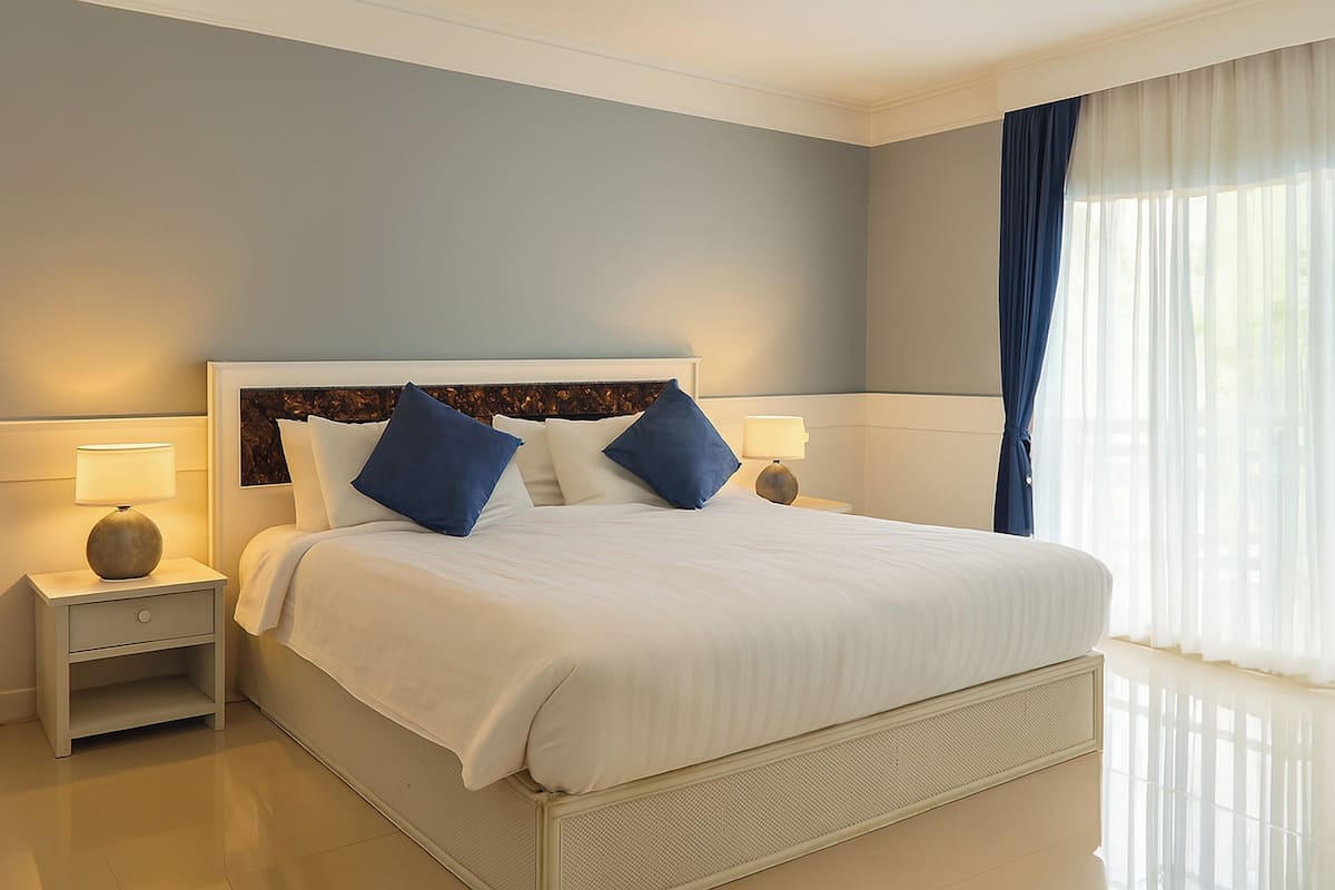 Una cama bien hecha con ropa de cama blanca y almohadas azules, una mesita de noche con lámpara y una ventana con cortinas translúcidas.