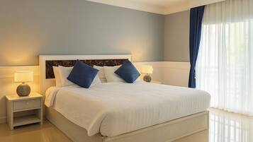 A neatly made bed with white bedding and blue pillows, a bedside table with a lamp, and a window with sheer curtains.