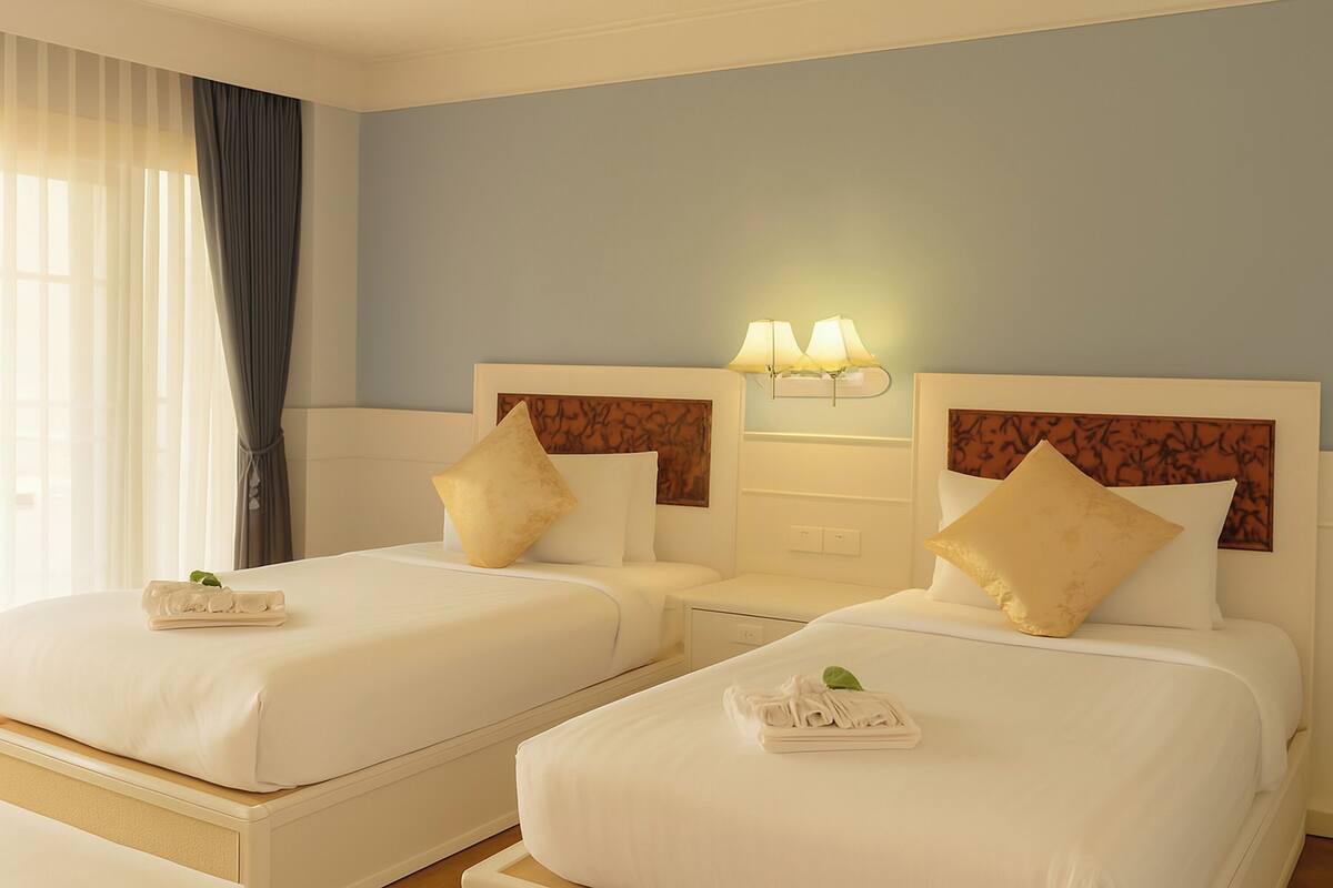 Habitación de hotel con dos camas, cada una con ropa blanca y cabeceros a juego. Hay dos lámparas de noche y una mesita entre las camas.