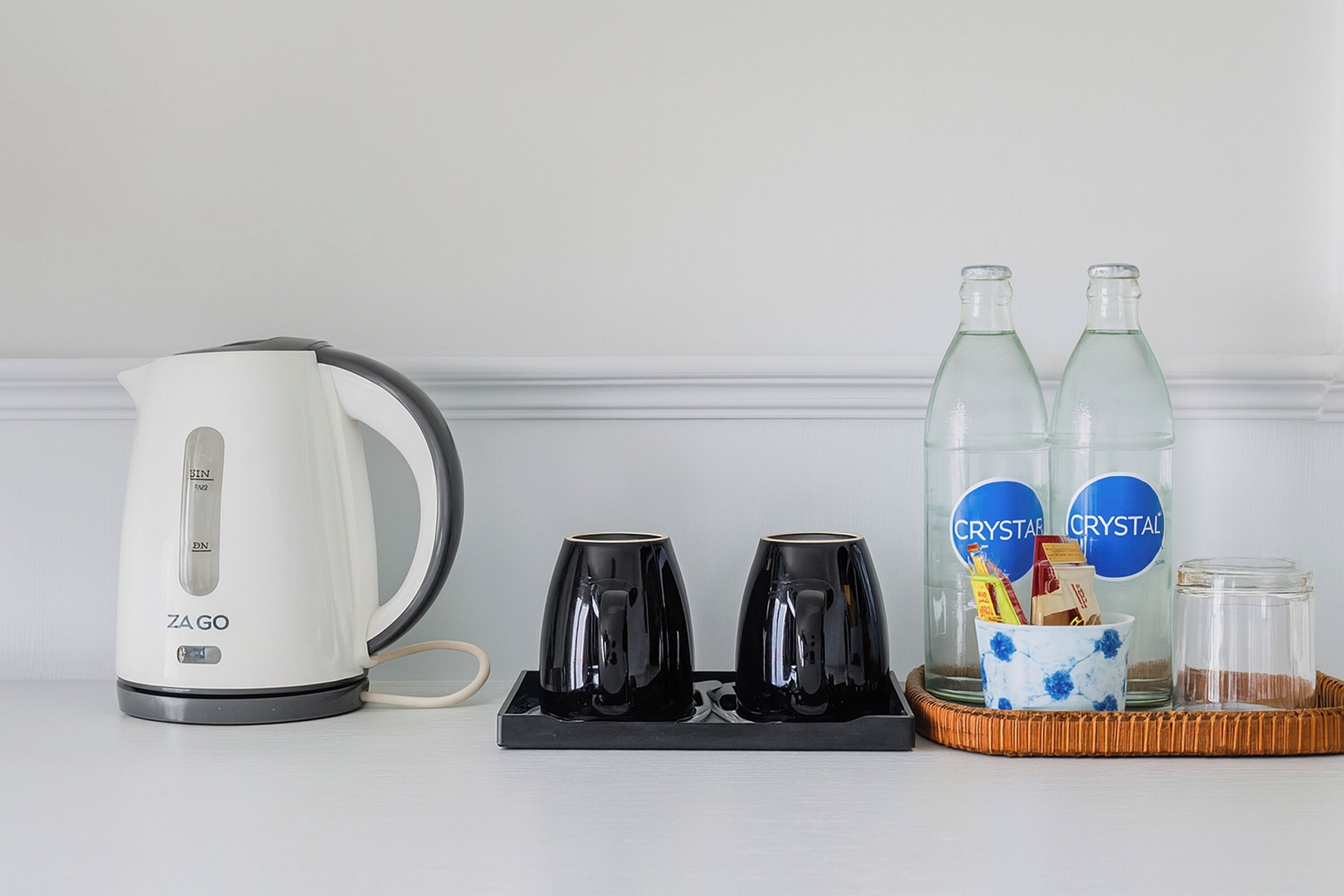 A kitchen setup with a ZAGO electric kettle, two black mugs, two Crystal water bottles, a small blue and white floral cup, and a glass jar.