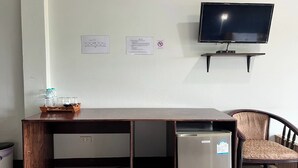 Una habitación con un escritorio de madera, un refrigerador pequeño, un televisor sobre un mueble y una silla.