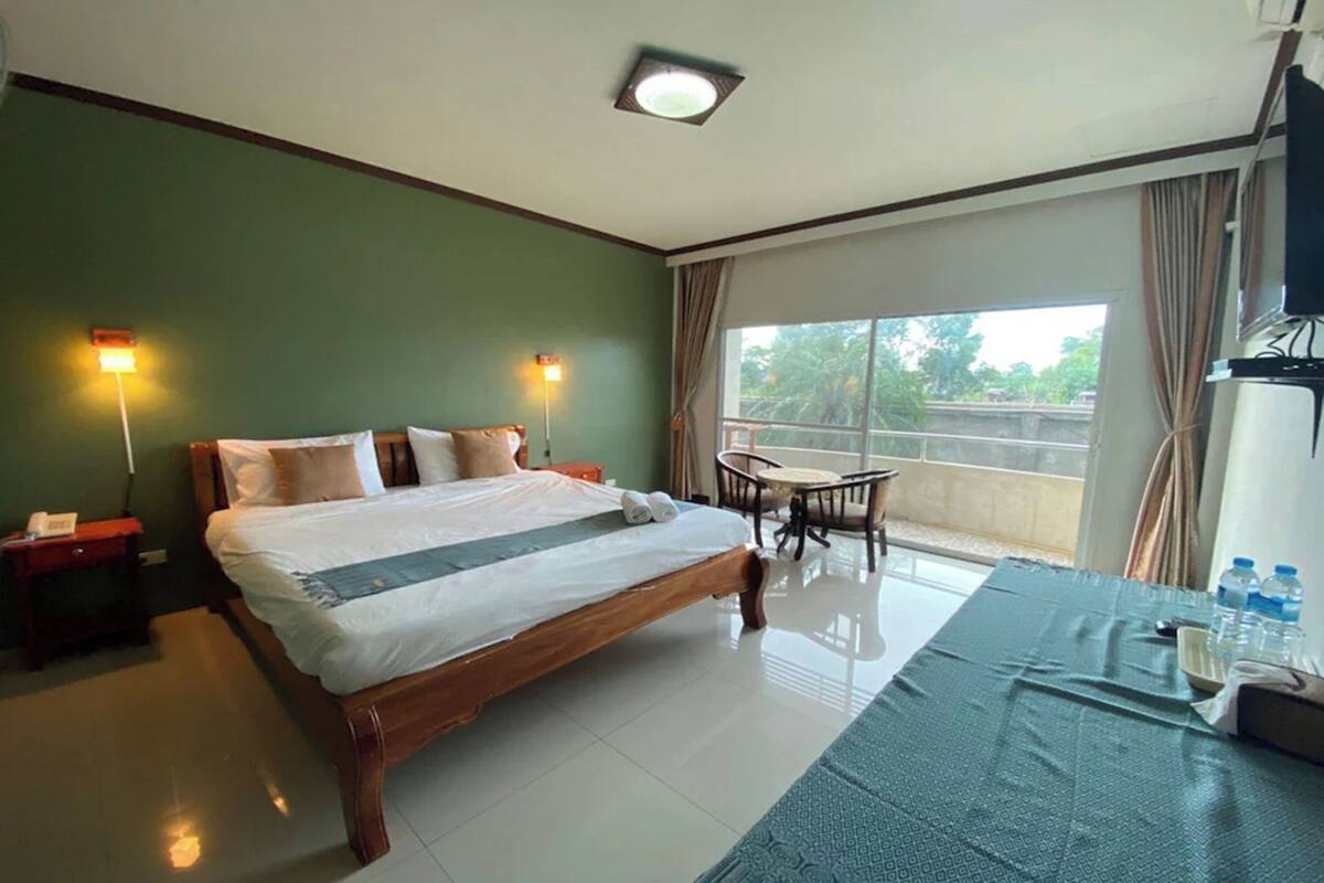 A hotel room with a large bed, bedside tables, a small table with chairs, a TV, and a balcony with curtains.