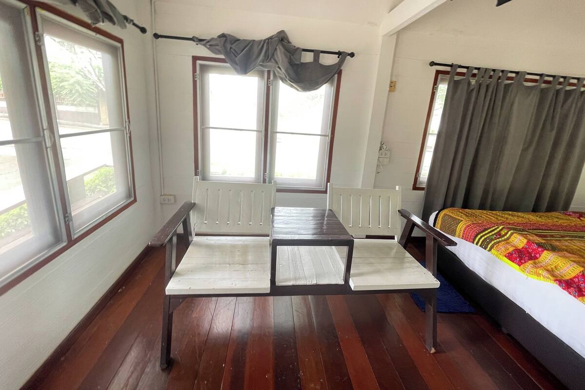 Une pièce comprenant un lit, une table et deux chaises. Il y a deux fenêtres avec des rideaux, un radiateur et une prise de courant au mur.