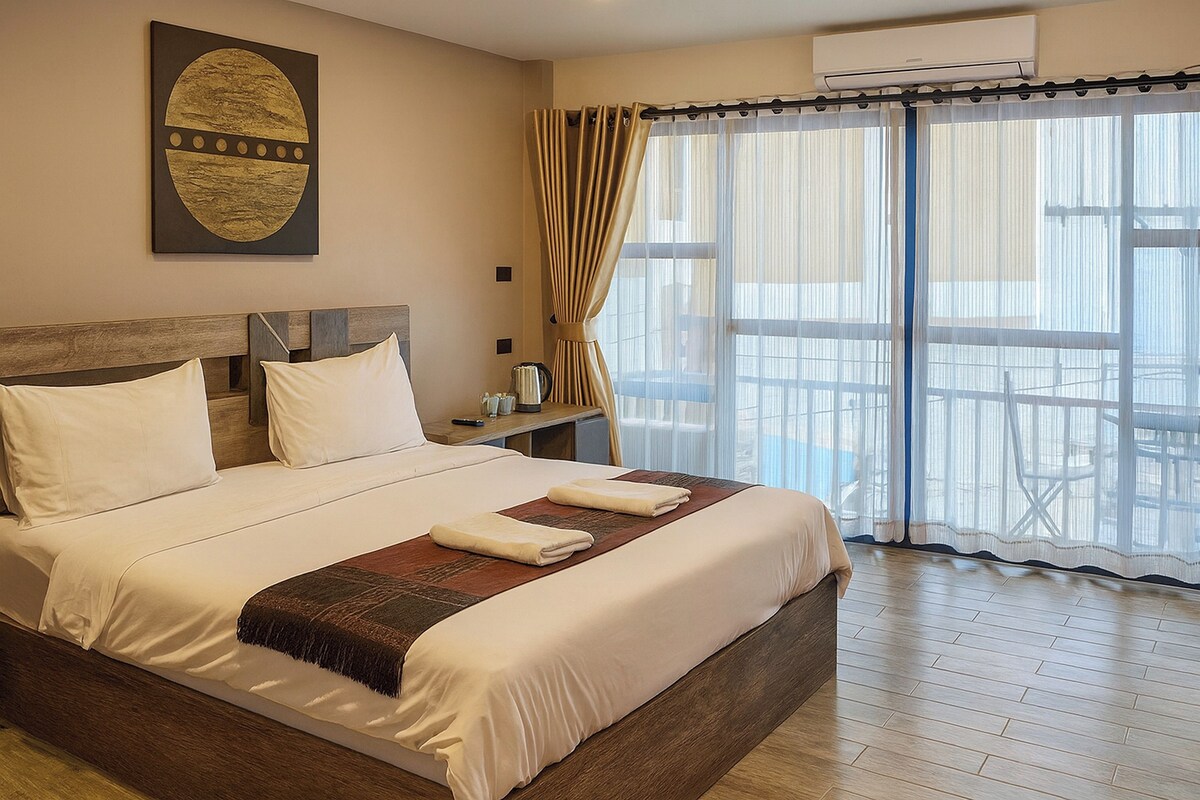 A neatly made bed with white linens and a brown throw blanket, a wooden headboard, and a large window with sheer curtains.