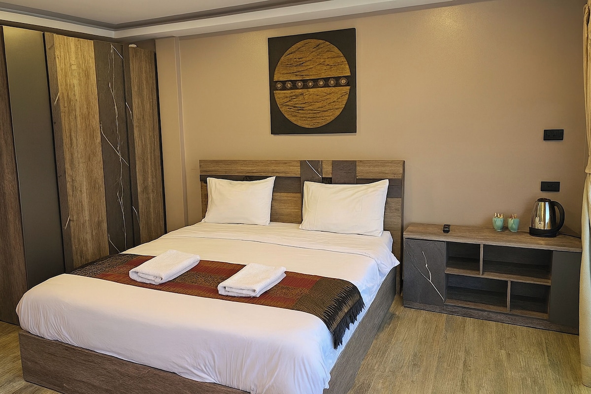 A neatly made bed with white linens and two towels, a wooden headboard, a wall-mounted artwork, a bedside table with a kettle, and a shelf unit.