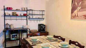 Een eetruimte met een houten tafel gedekt voor vier personen, omringd door stoelen. Op de tafel staan een koffiezetapparaat en een waterkoker, en tegen de muur bevindt zich een plank met keukengerei.