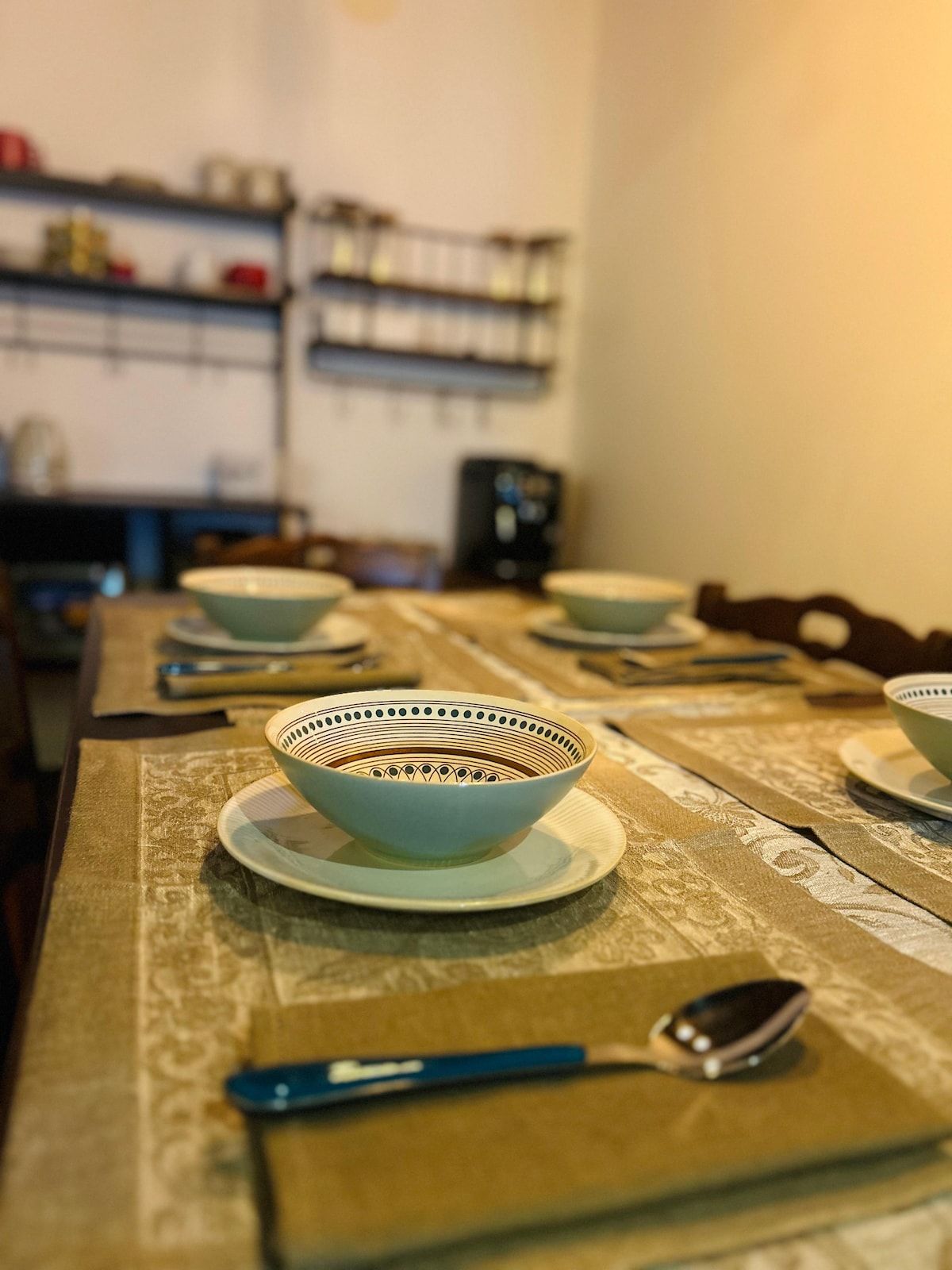 A dining table with bowls and a spoon, a patterned tablecloth, and shelves in the background.