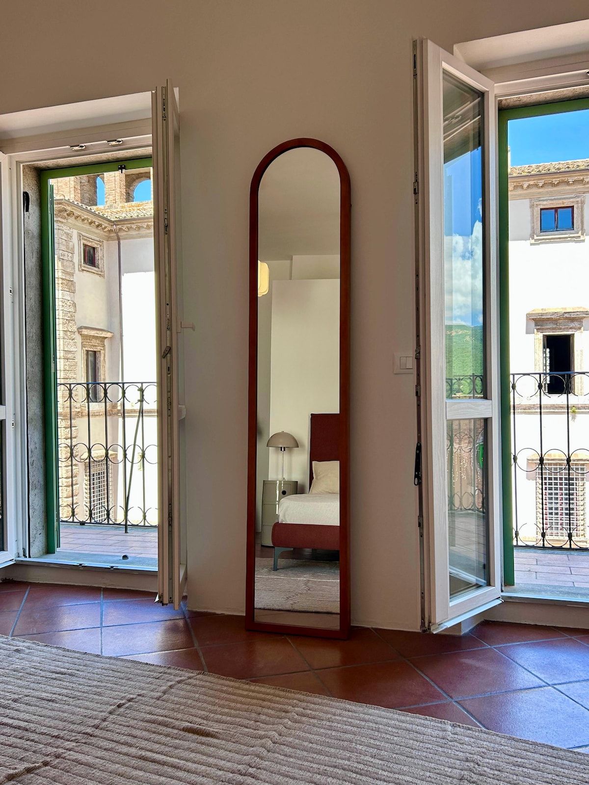 Une pièce avec un grand miroir, un lit, une table de chevet et une vue sur un immeuble à travers une porte vitrée.