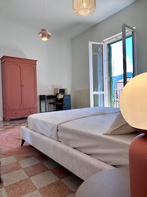A bedroom with a bed, a desk, a chair, a wardrobe, and a view through an open door.