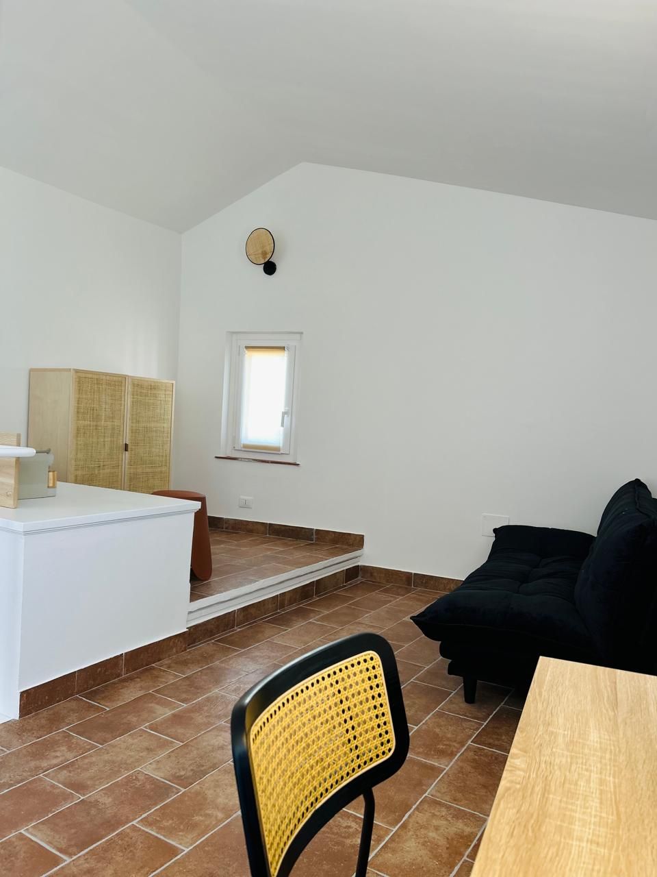 Una stanza con un muro bianco, una finestra, un divano nero, un tavolo di legno, una sedia in vimini e un piccolo armadietto bianco.